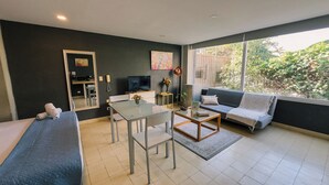 Een moderne woonkamer met een bank, een eettafel met stoelen, een televisie en een groot raam met uitzicht op groen.