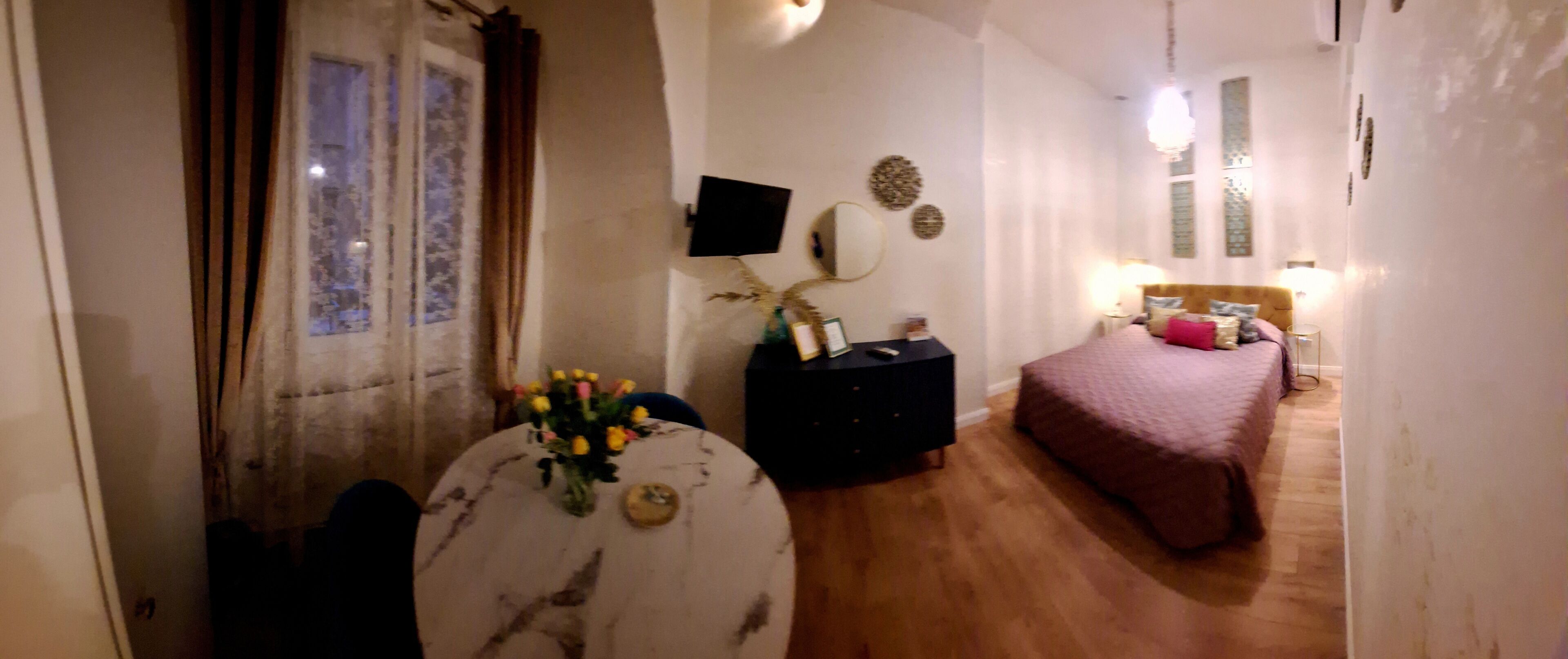 Een slaapkamer met een bed, nachtkastje, lamp en een tafeltje met een bloemenvaas.