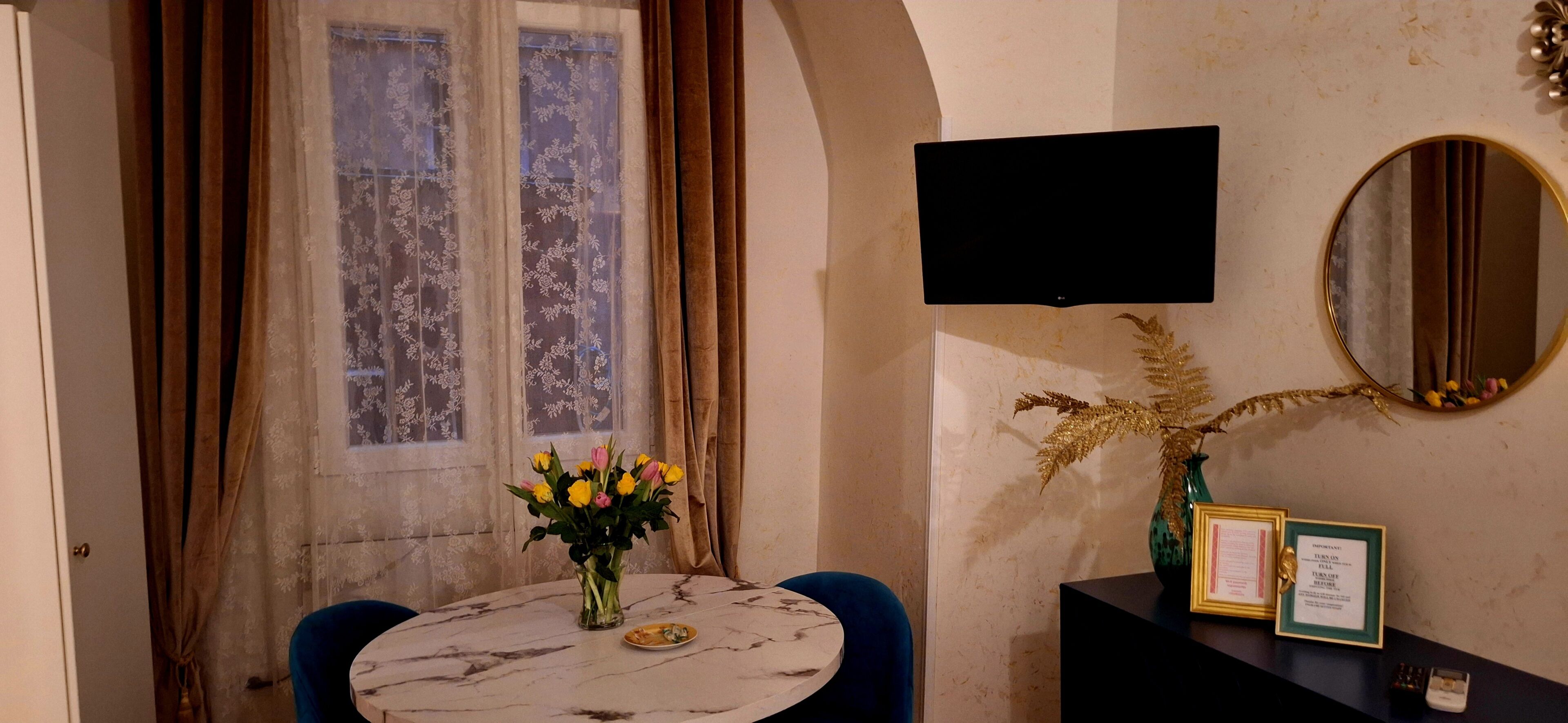 A room with a round table, a vase of flowers, a mirror, and a framed certificate on a black stand.