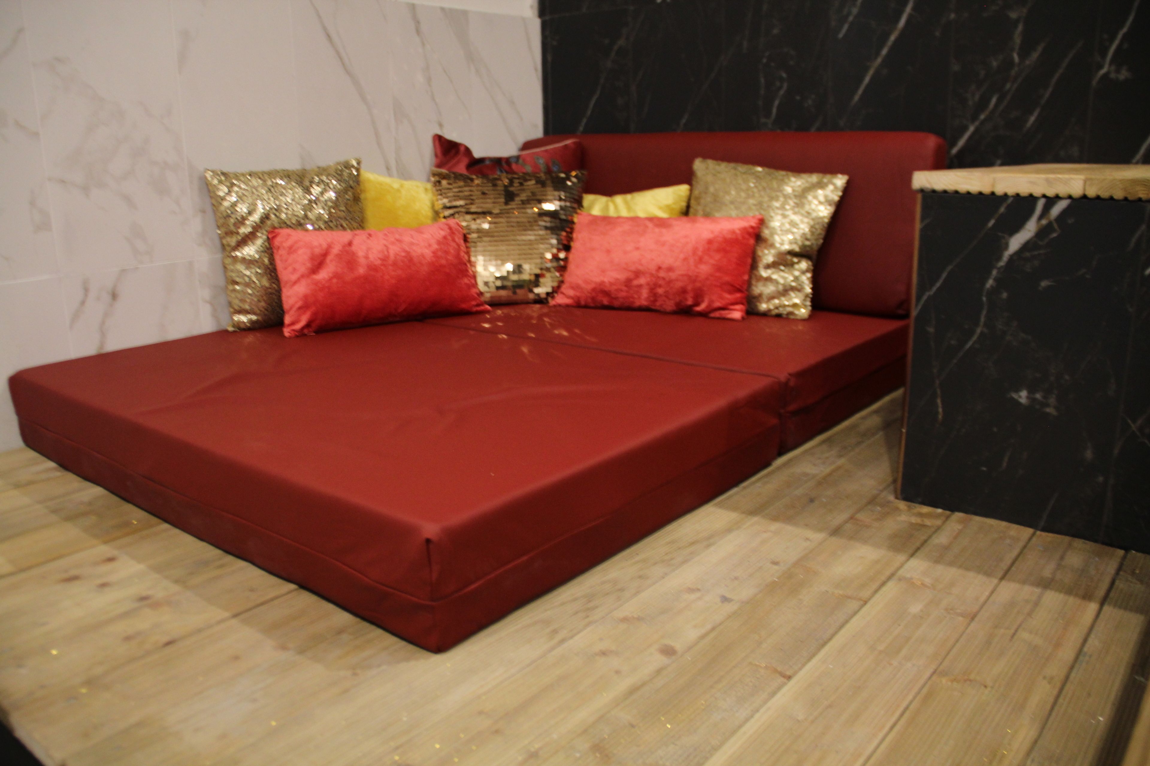 A red sofa bed with multiple cushions in a room with a wooden floor and a marble wall.