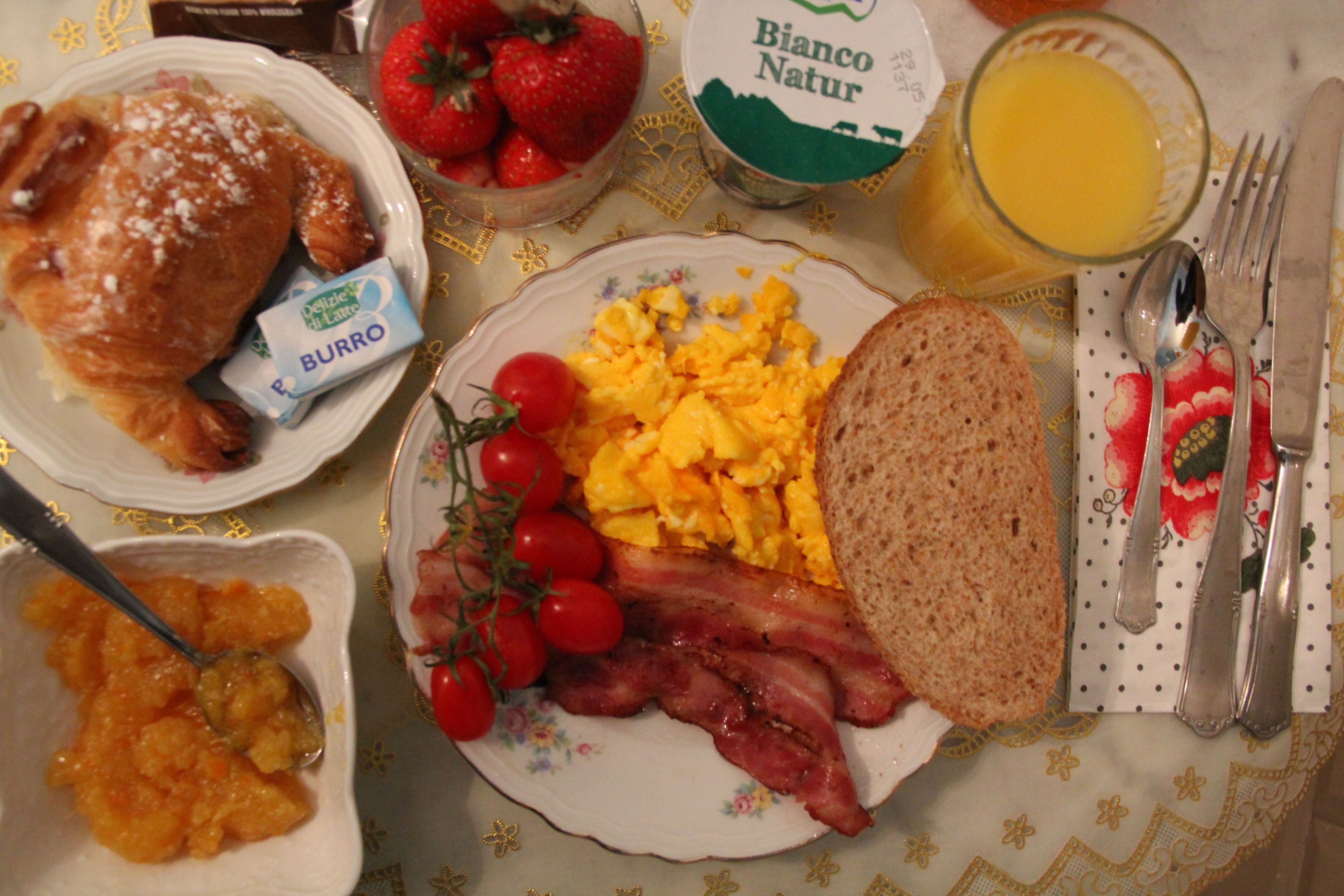 Een ontbijttafel met roerei, spek, toast, aardbeien en een pak melk.