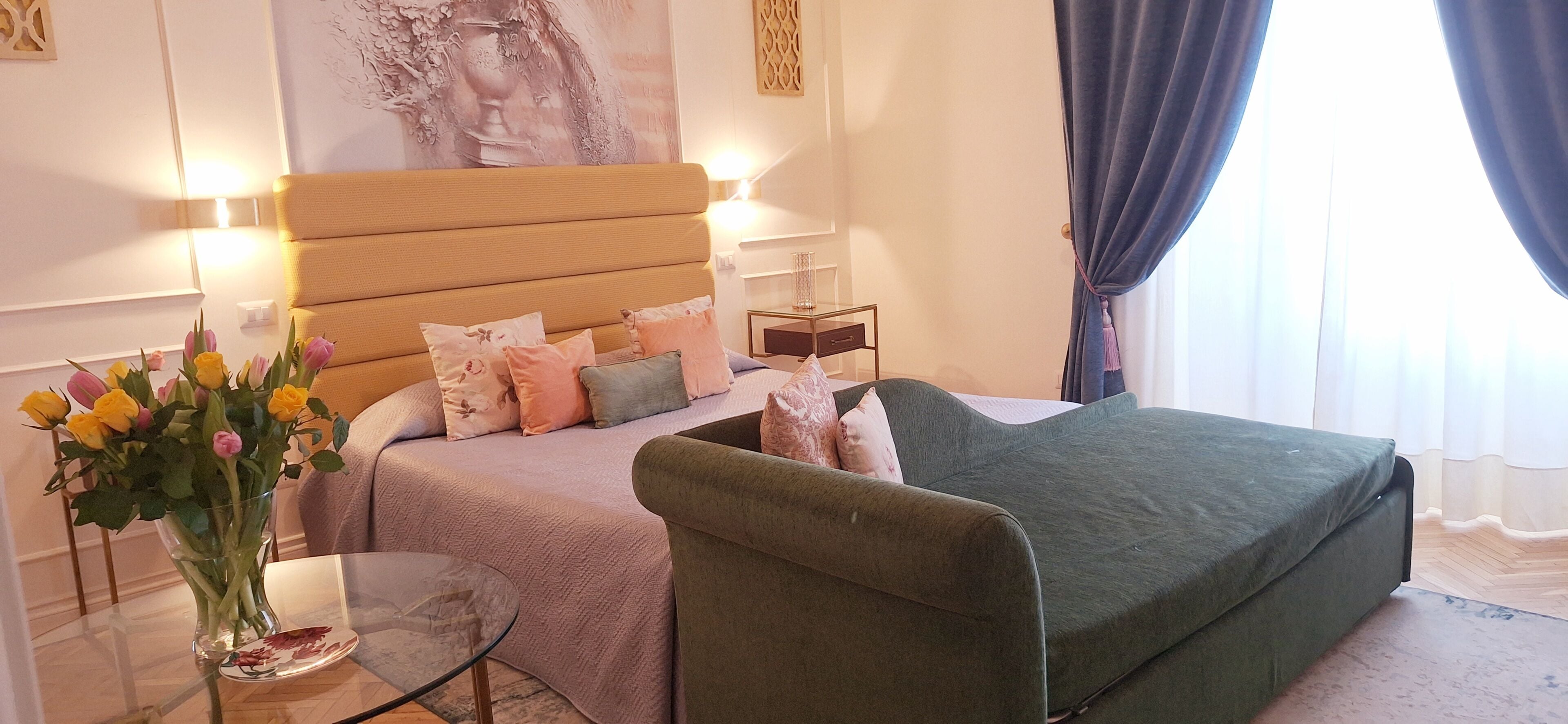 Un dormitorio con cama, una silla, una mesita de noche, un florero con flores y una lámpara de pared.