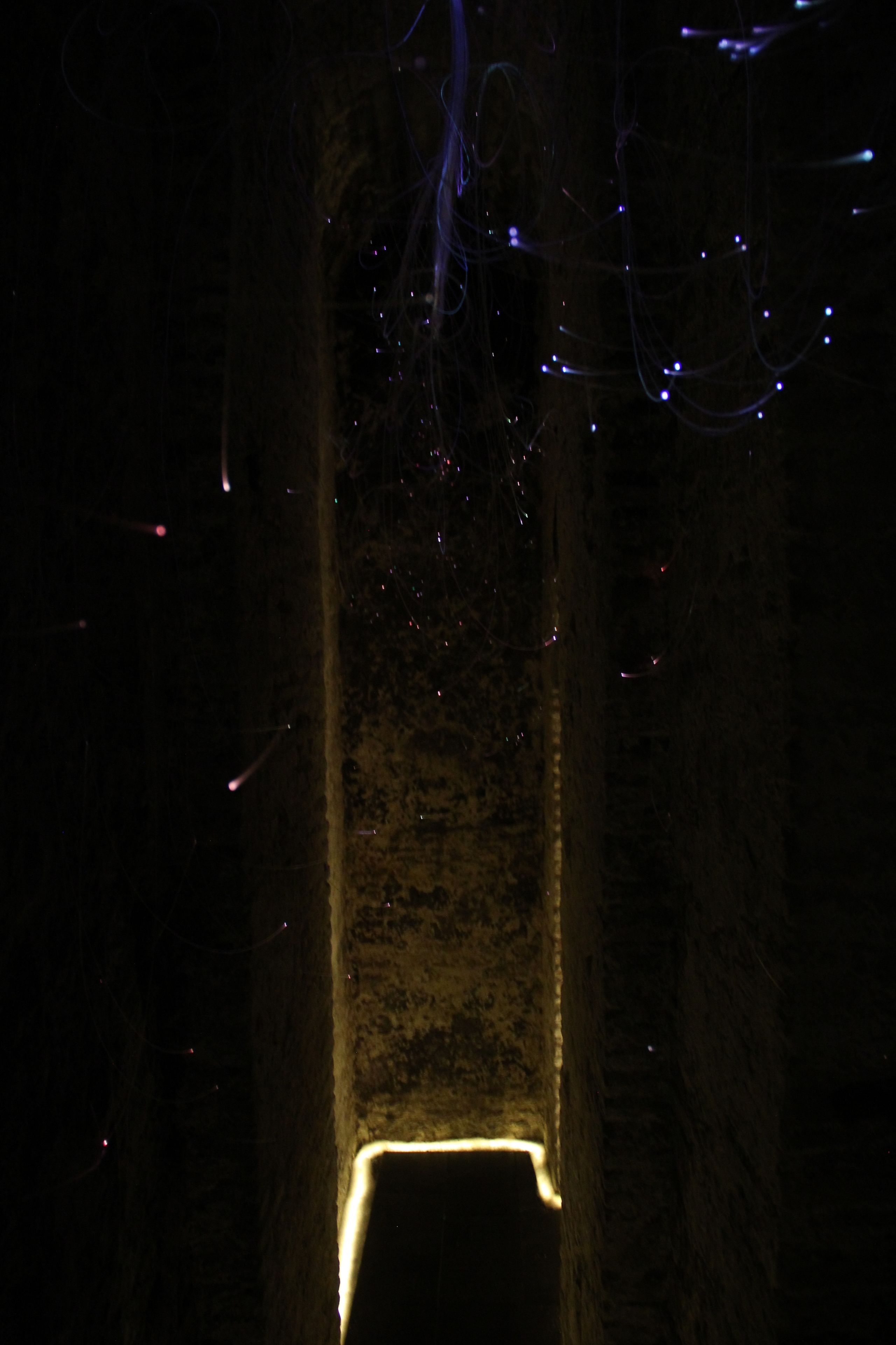 A dark, narrow passageway with a light at the end, surrounded by rough stone walls.