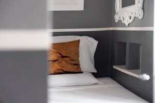 A neatly made bed with white and brown bedding, a white pillow, and a small white shelf against a grey wall.