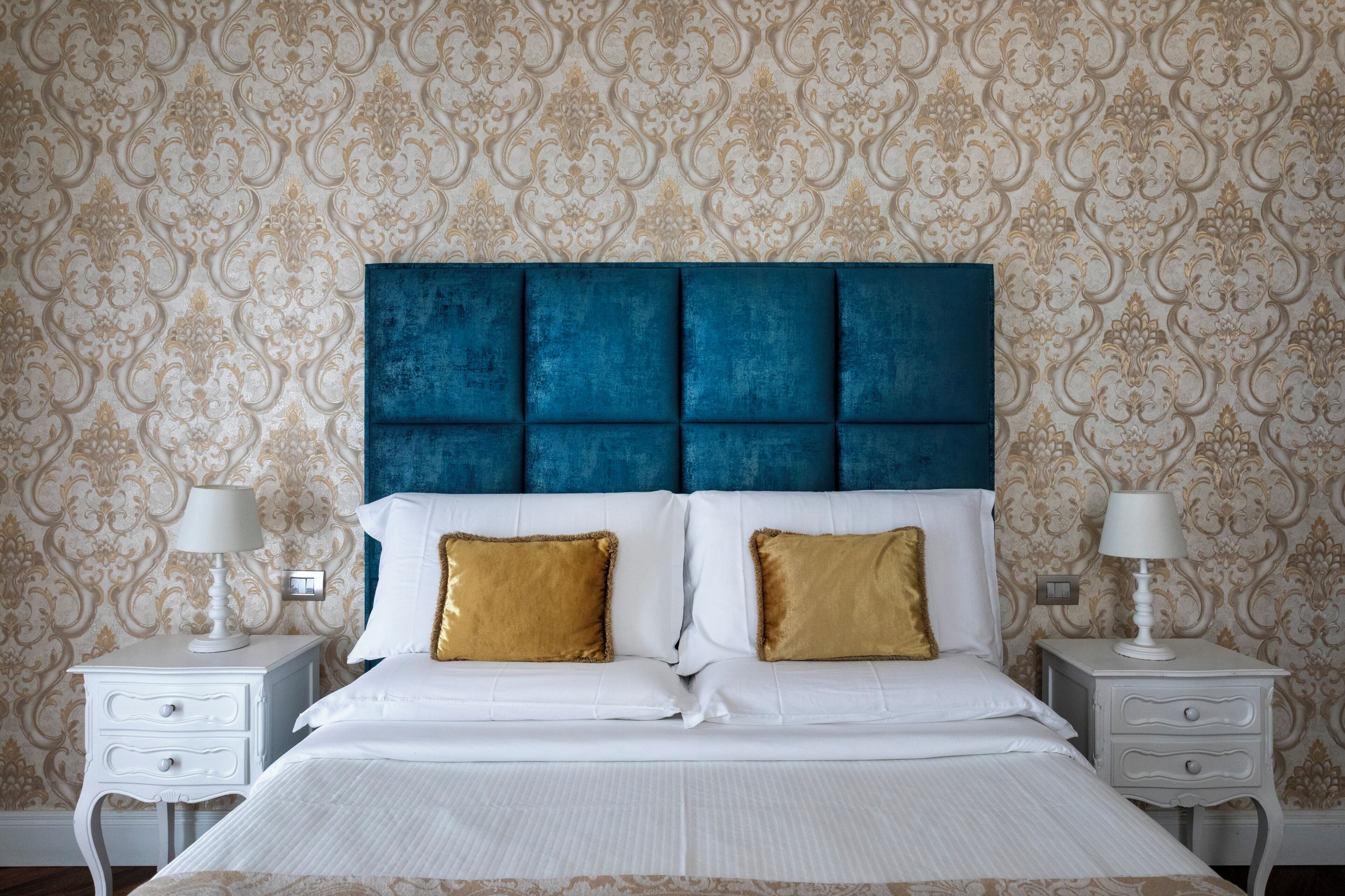 Un dormitorio con cabecera capitonada, ropa de cama blanca, dos almohadas doradas decorativas y mesitas de noche con lámparas.