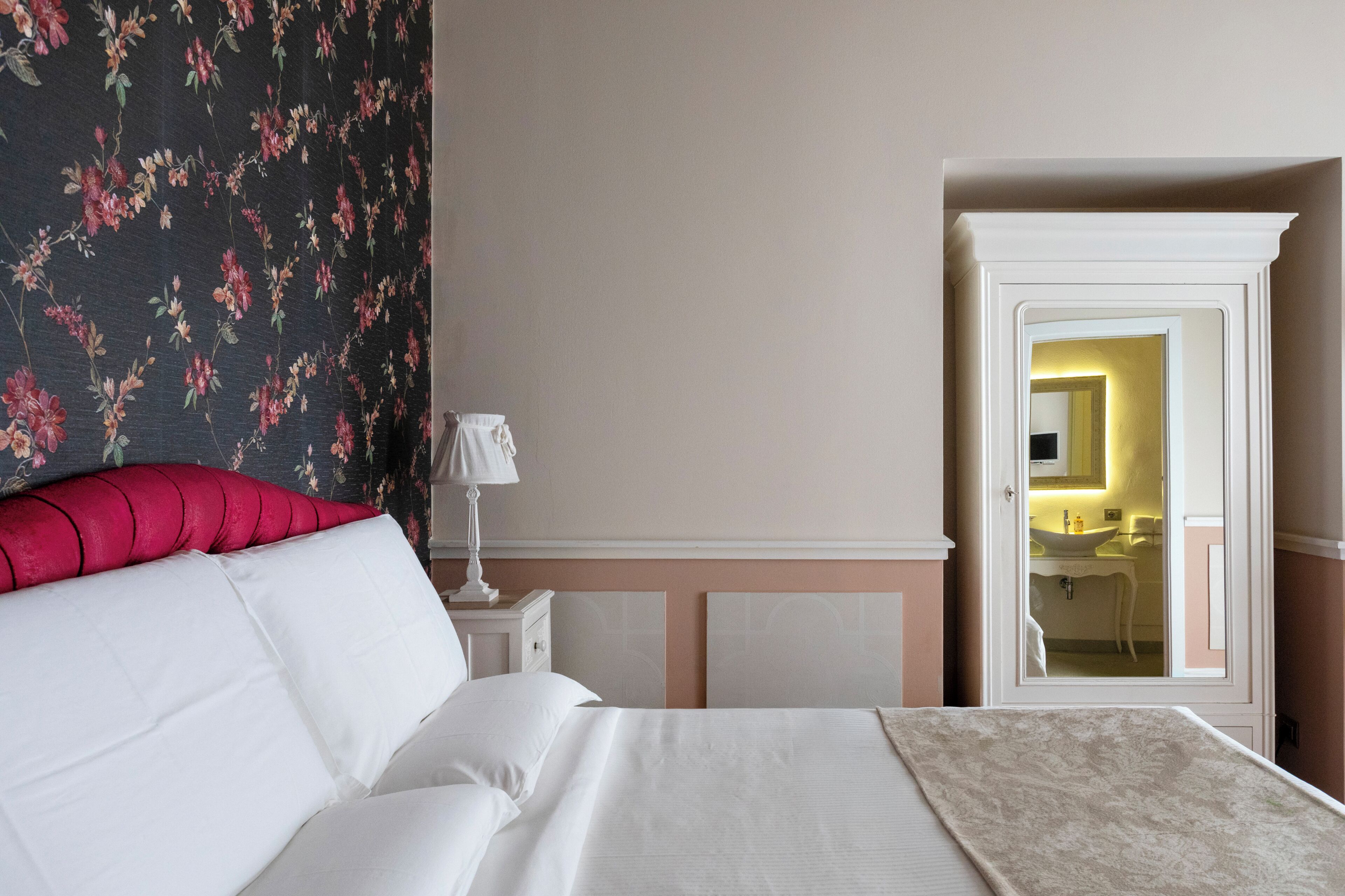 A bedroom with a bed, a nightstand, a lamp, a floral wallpaper, and a view of a bathroom through an open door.