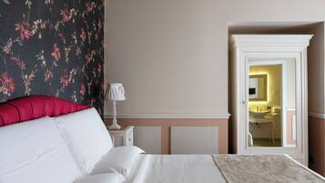 A bedroom with a bed, a nightstand, a lamp, a floral wallpaper, and a view of a bathroom through an open door.