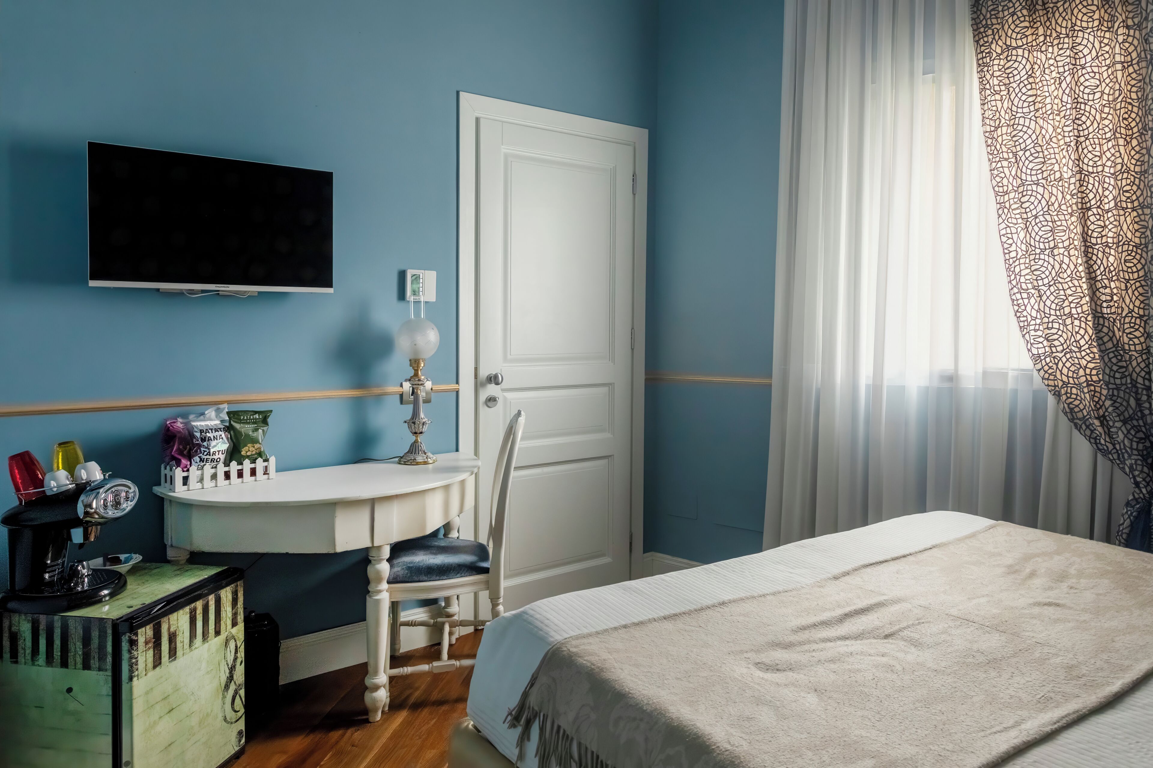 Une chambre à coucher avec un lit, un bureau et une chaise, un téléviseur fixé au mur, une porte et une fenêtre avec des rideaux.
