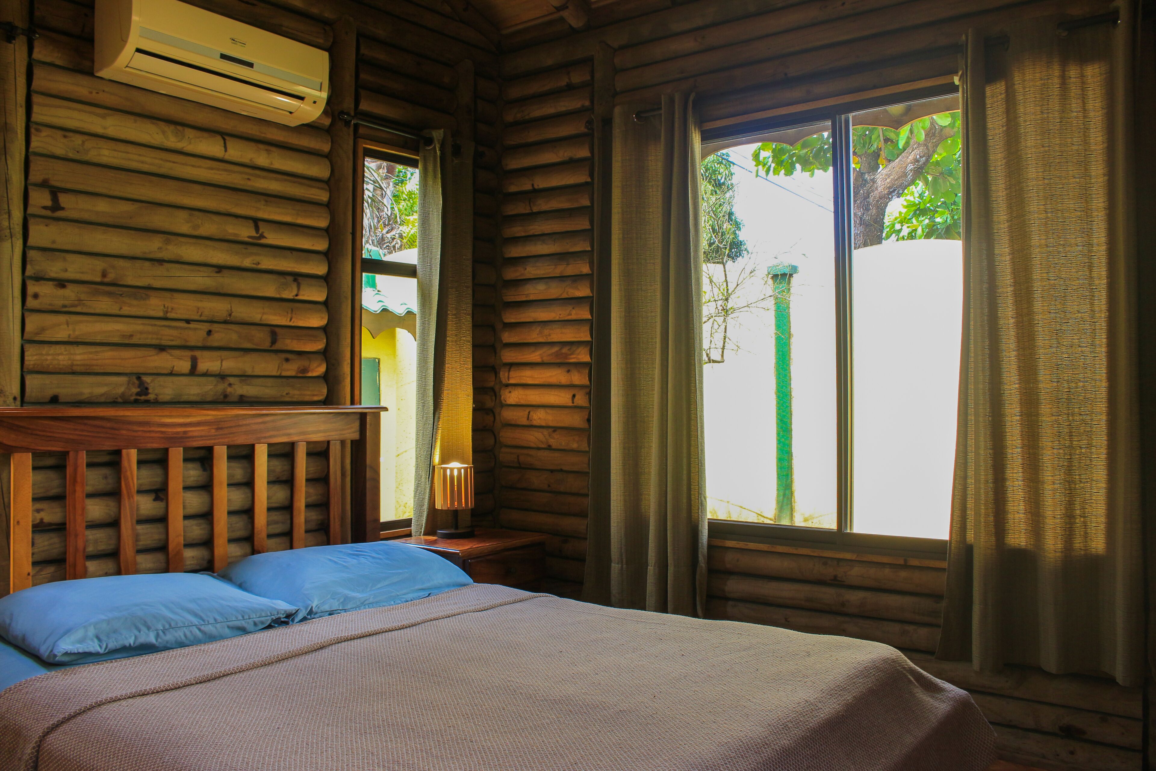 A wooden cabin room with a bed, a nightstand, and a window with curtains.