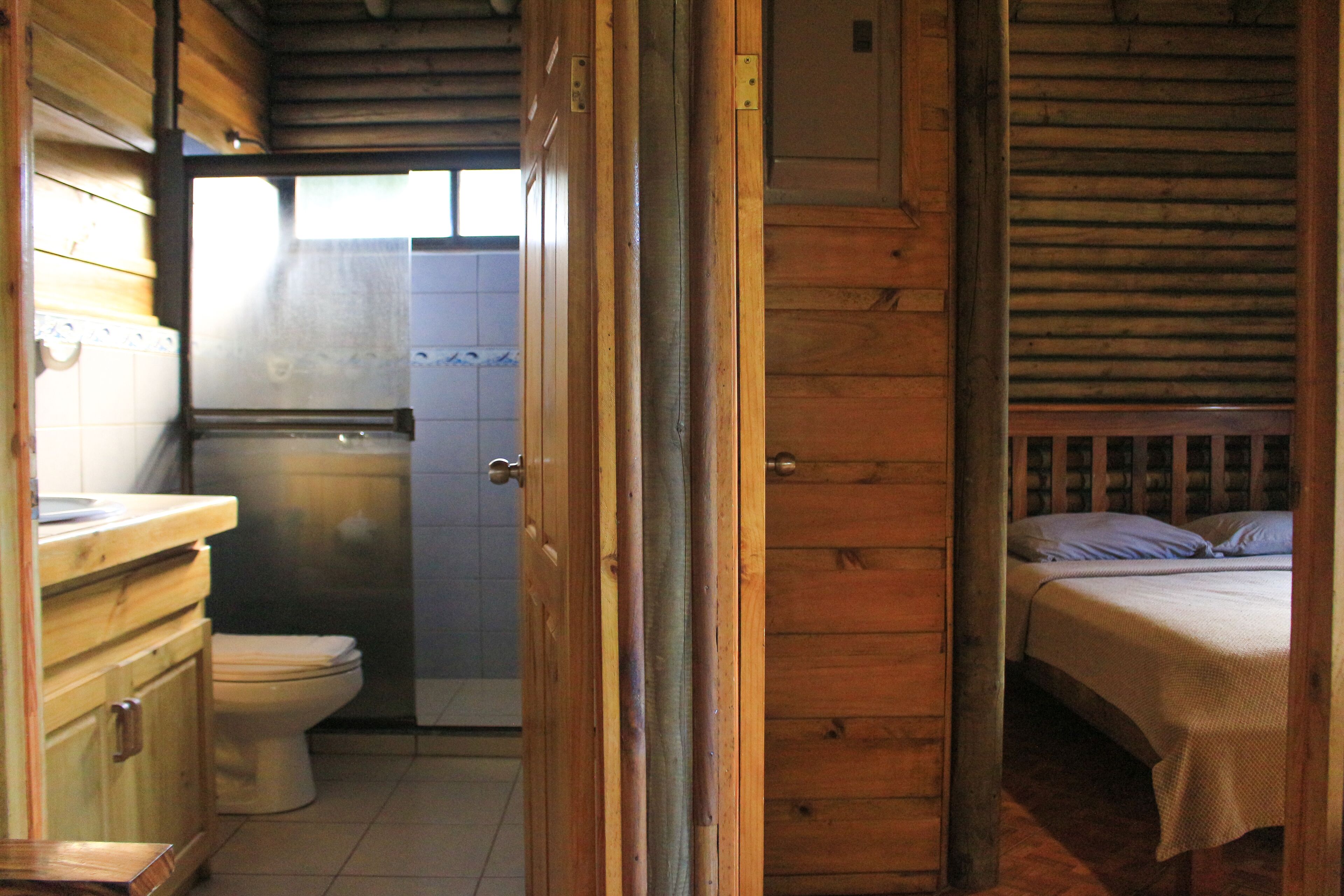 A wooden cabin interior with a wooden door leading to a bathroom, a toilet, and a bed with a wooden headboard.