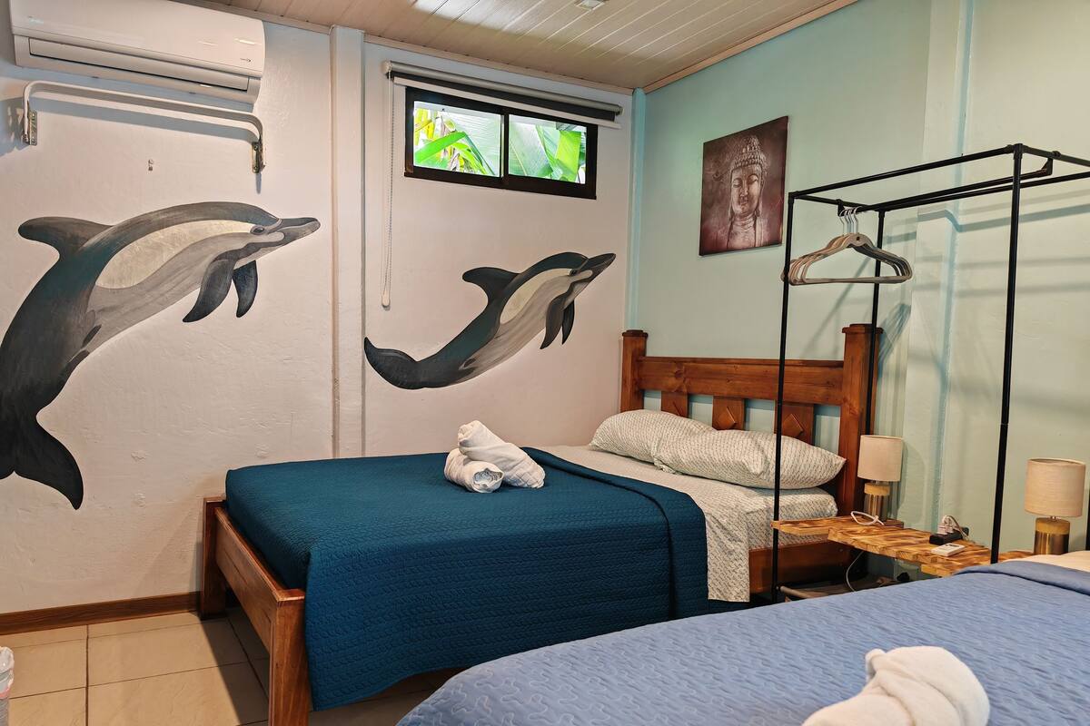 A room with two beds, one with a blue bedspread and the other with a white one. There is a wooden headboard, a window with a view of greenery, and a wall with dolphin murals.