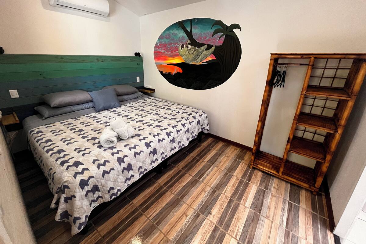 A bedroom with a bed, a wall-mounted air conditioner, a wooden headboard, a wall art of a tree, and a wooden wardrobe.