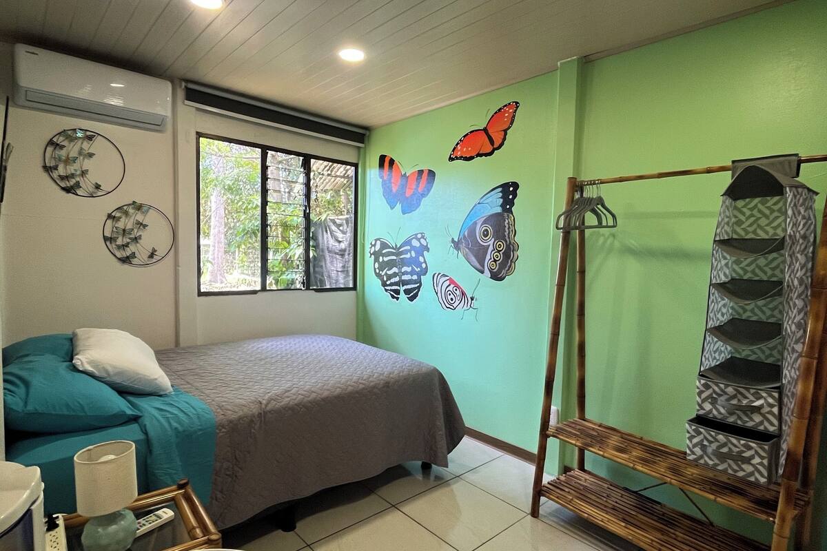 A room with a bed, a bedside table with a lamp, a wall-mounted air conditioner, a window, and a wall with butterfly decorations.