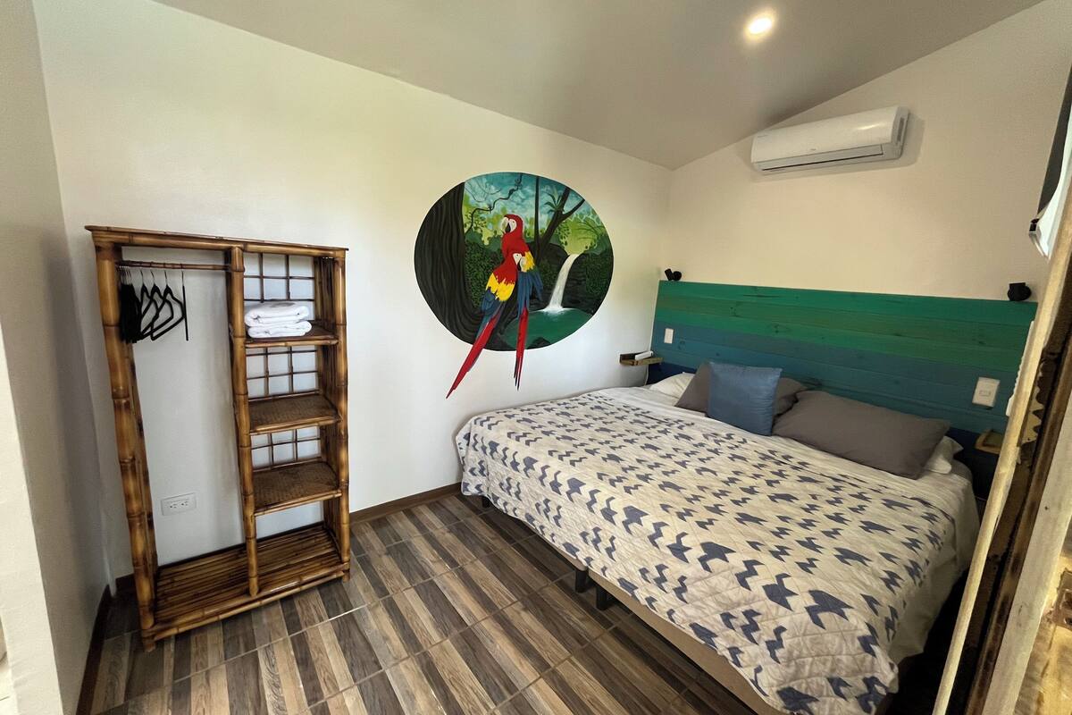 A bedroom with a bed, a wall-mounted air conditioner, a wall art of a parrot, and a wooden shelf unit.