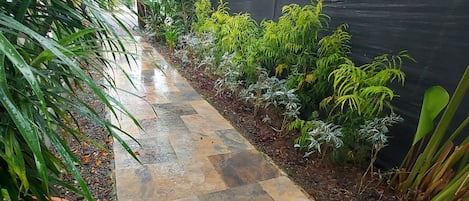 A pathway with stone tiles, surrounded by various plants and a black fence.
