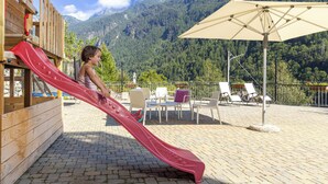 A wooden play structure with a red slide, outdoor seating, and a view of a mountain range. - Hotel Biancospino (Lanzada)