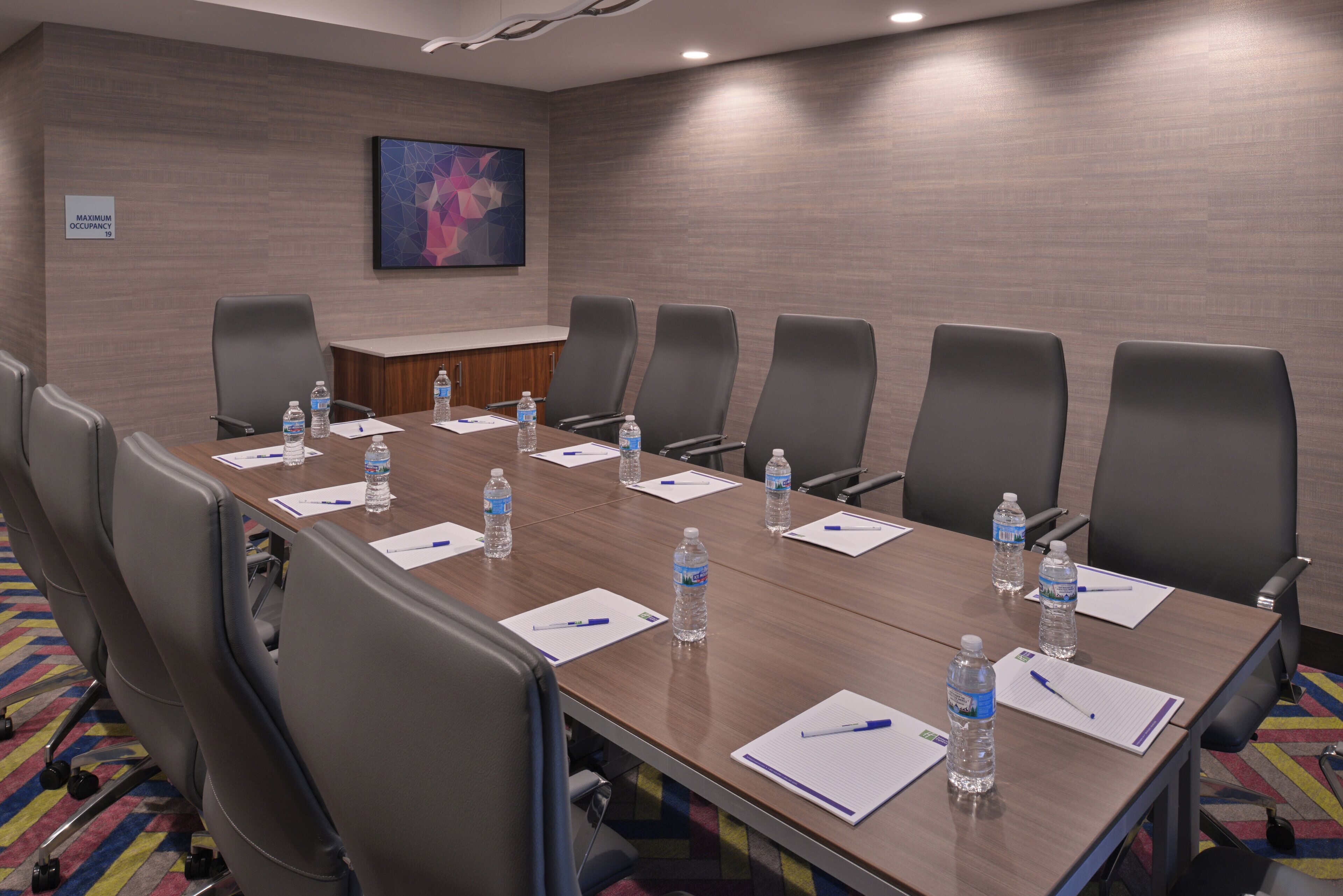 Uma sala de conferência com uma mesa comprida, oito cadeiras, garrafas de água e papéis sobre a mesa.