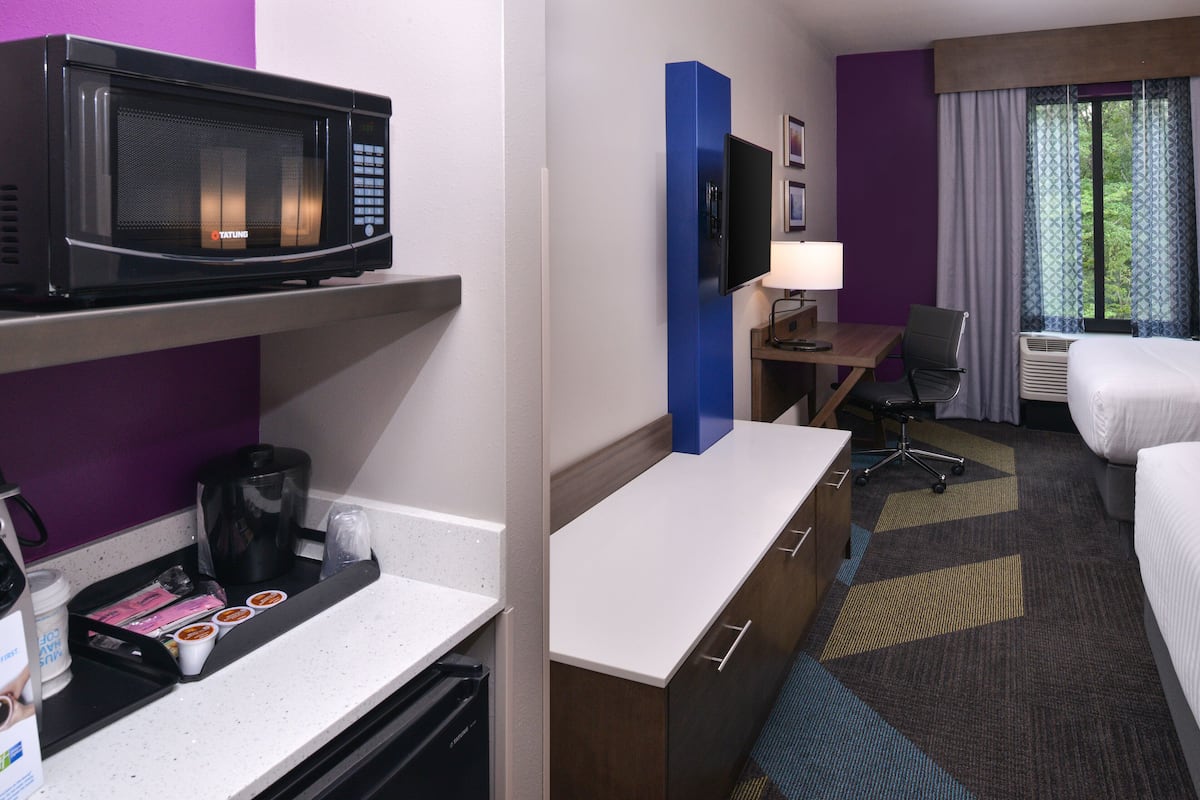 Habitación de hotel con microondas, cafetera y una pequeña mesa con una silla. Incluye una cama, un escritorio con lámpara y una ventana con cortinas.