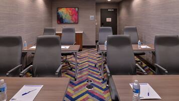 Un salón de conferencias con alfombra estampada en forma de rombos, sillas grises, mesas de madera y botellas de agua sobre las mesas.