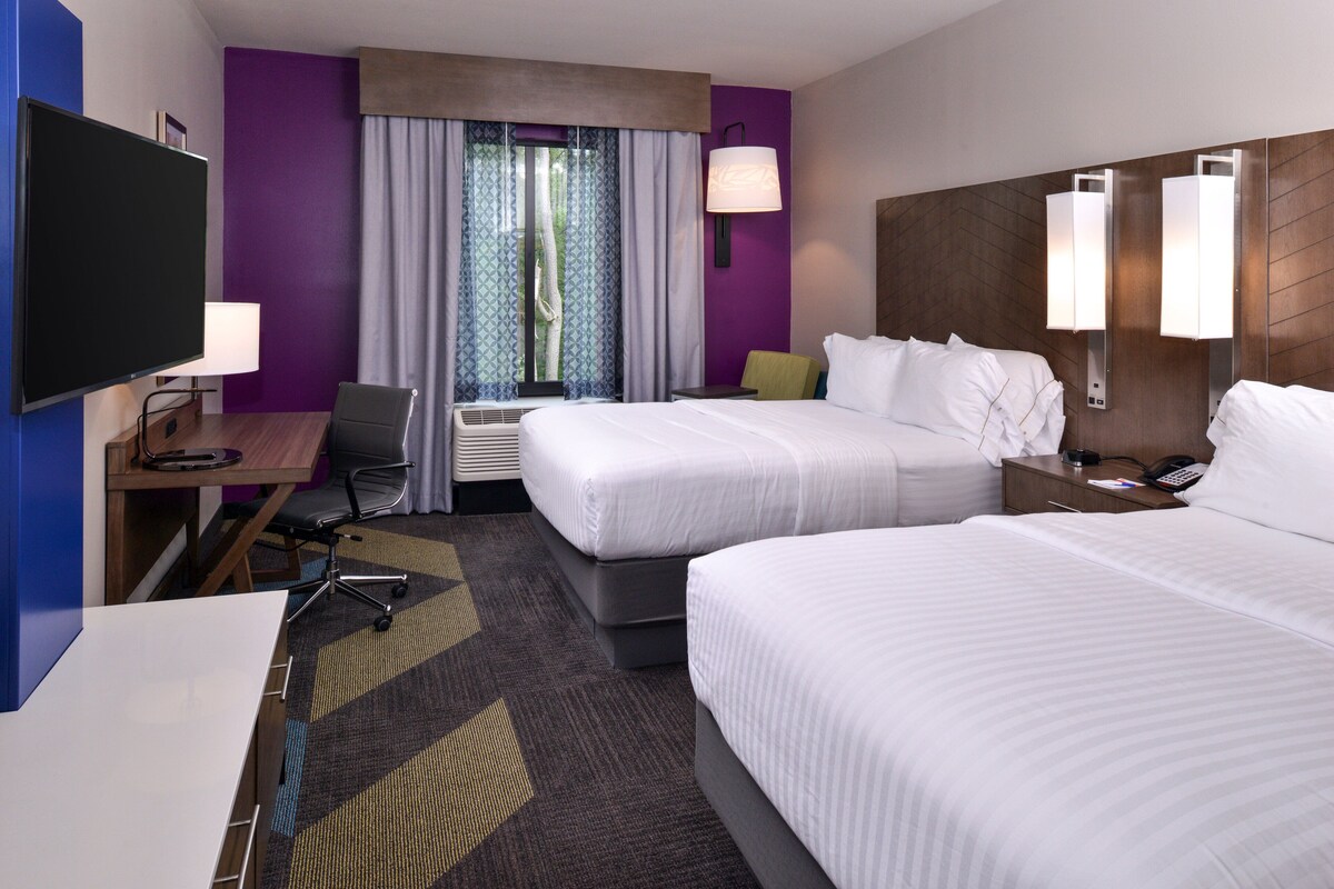 A hotel room with two beds, a desk, a TV, and a window with curtains.