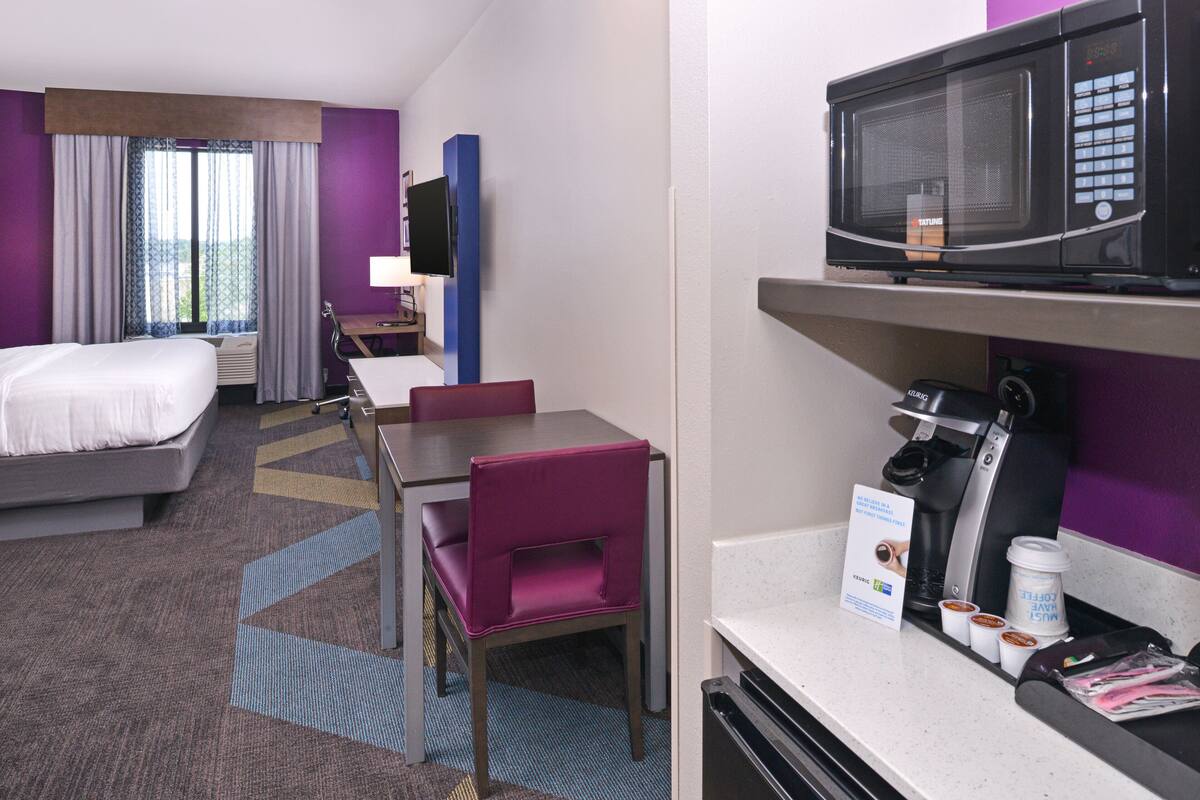 Una habitación de hotel con cama, escritorio con silla, microondas, cafetera y televisor.