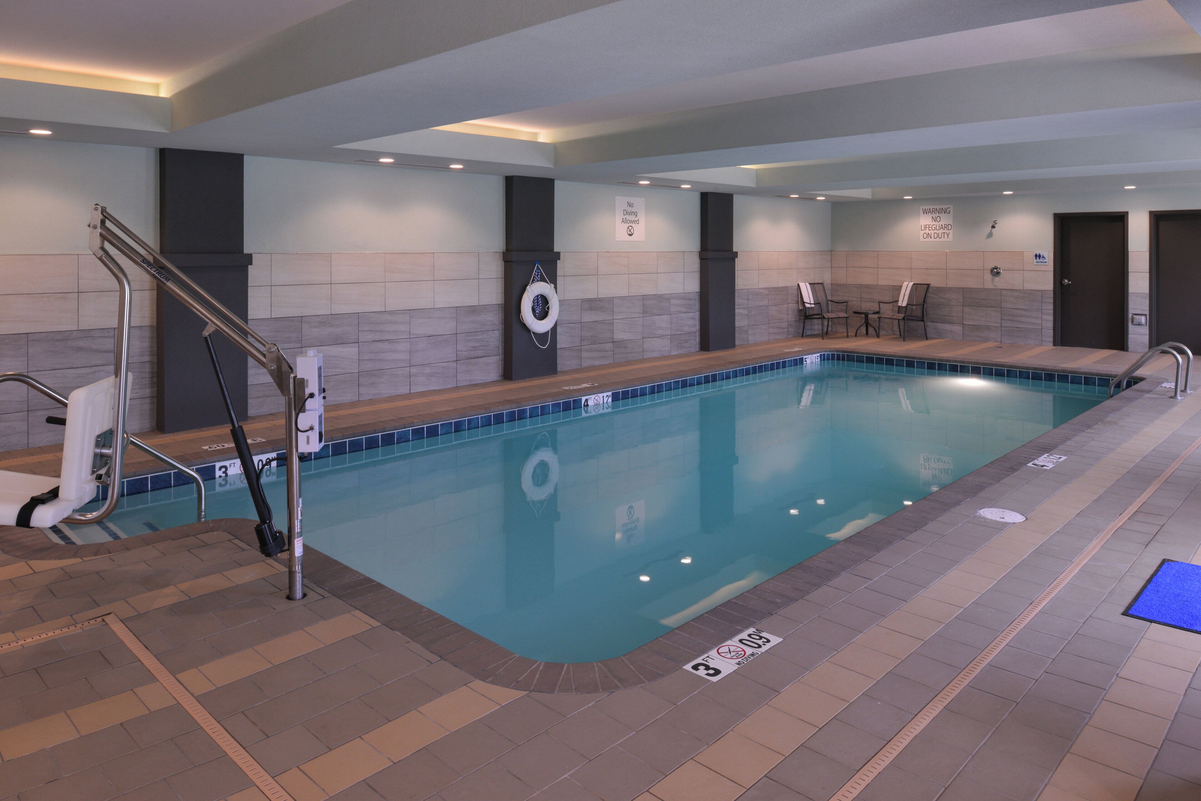 Una piscina interior con una silla de salvavidas, un letrero que indica que no se permite bucear y un aviso que indica ausencia de salvavidas.