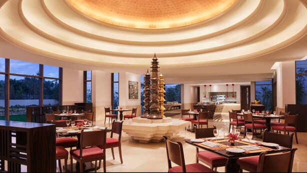 Un comedor amplio con varias mesas y sillas, un columnario decorativo en el centro y ventanas grandes que ofrecen vistas al exterior.
