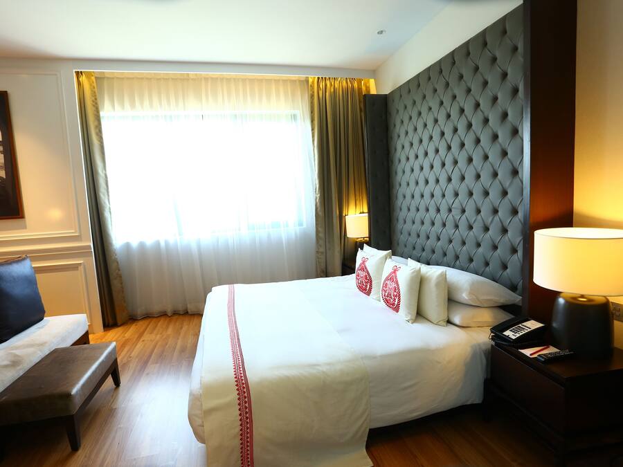 Habitación de hotel con una cama grande, un banco, una mesita de noche, una lámpara y vistas al exterior a través de una ventana con cortinas.