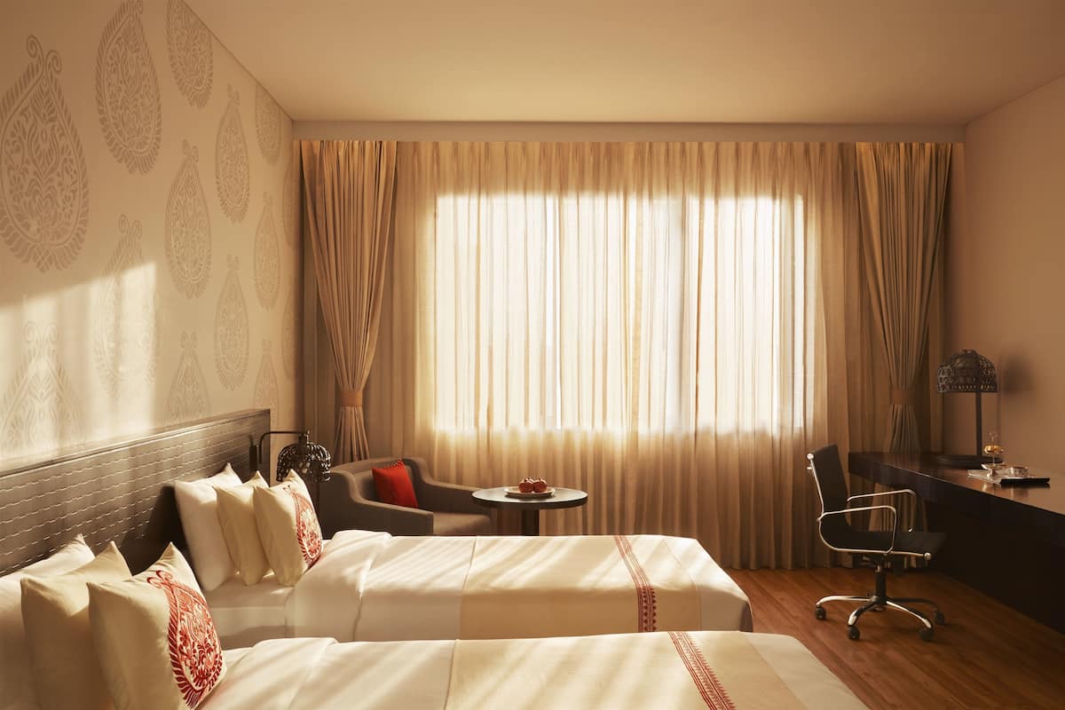 A hotel room with two beds, a desk, a chair, and a small table with a red cushion.