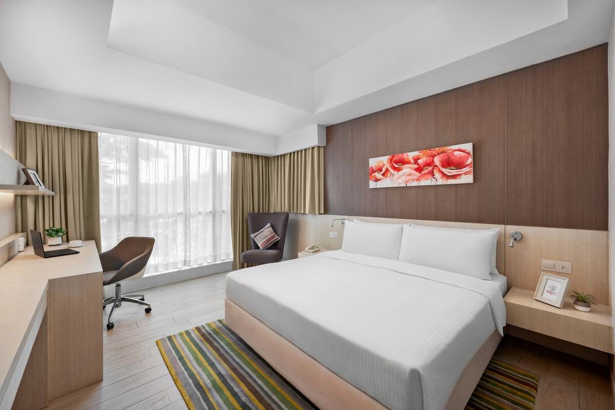 Habitació d'hotel moderna amb un llit gran, una taula de treball amb cadira, una taula petita i una finestra gran amb cortines.
