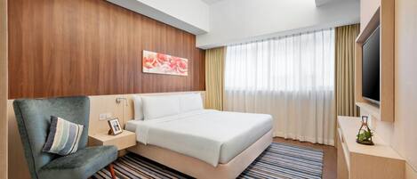 Una habitación de hotel moderna con una cama, una zona de estar con una silla, un televisor y un ventanal con cortinas.