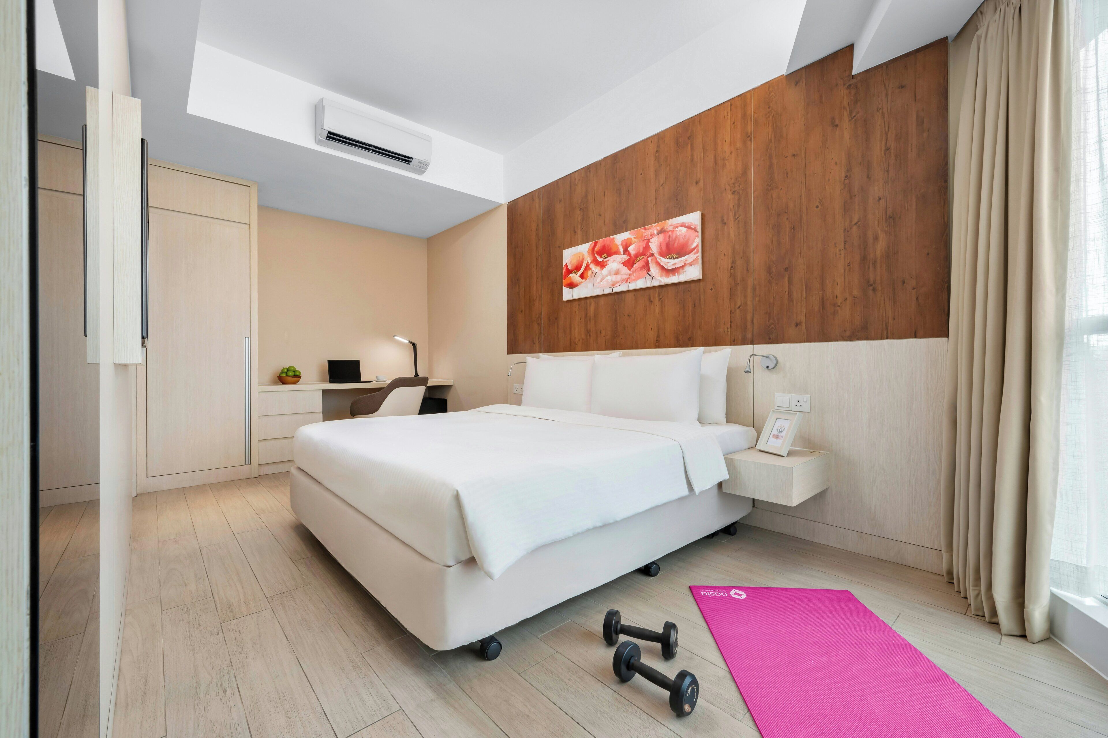 A modern hotel room with a bed, a desk, a chair, a small plant, a picture frame, and a pink exercise mat.
