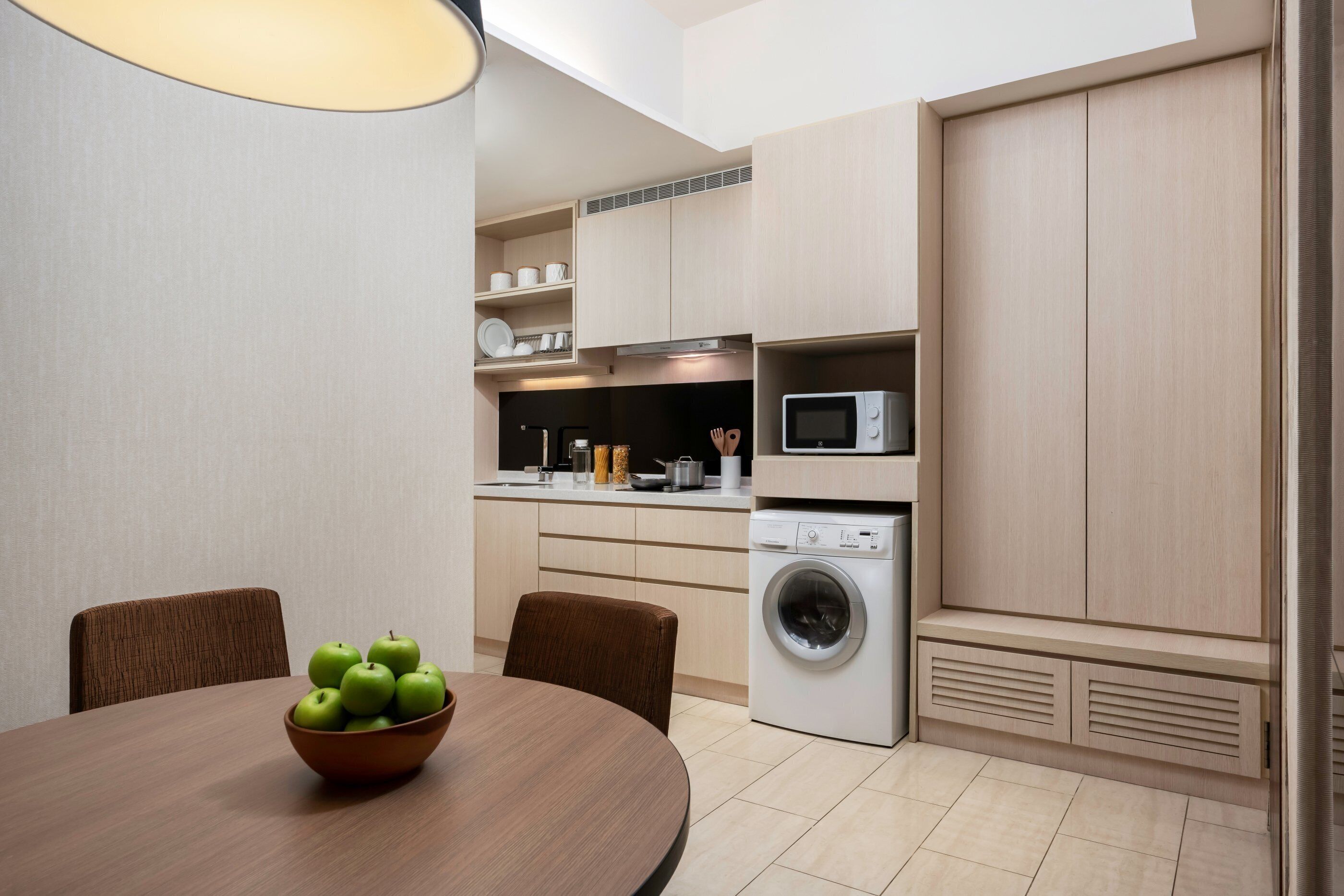 A modern kitchen with a dining table, chairs, a bowl of green apples, a microwave, and a washing machine.