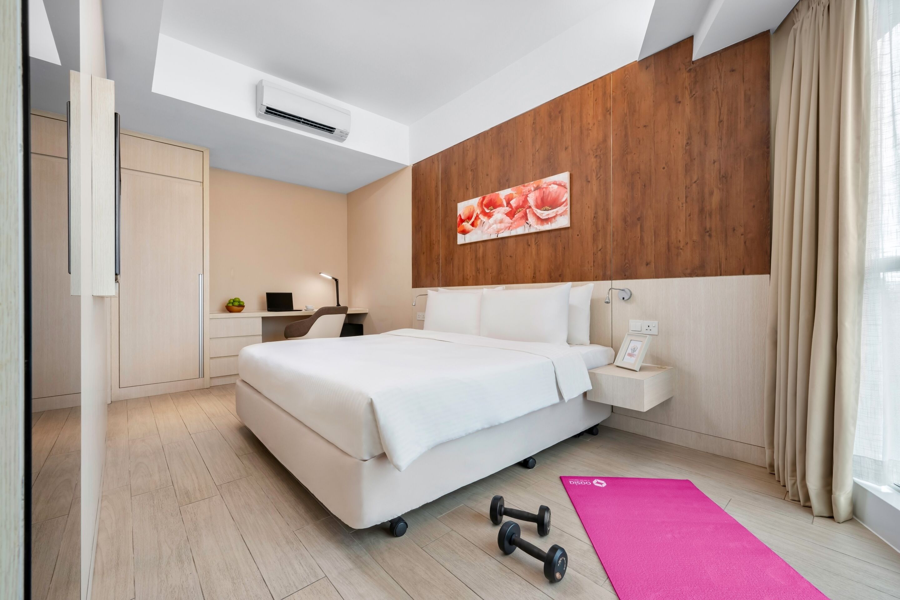 A modern hotel room with a large bed, a desk with a lamp, a small plant, and a workout mat.