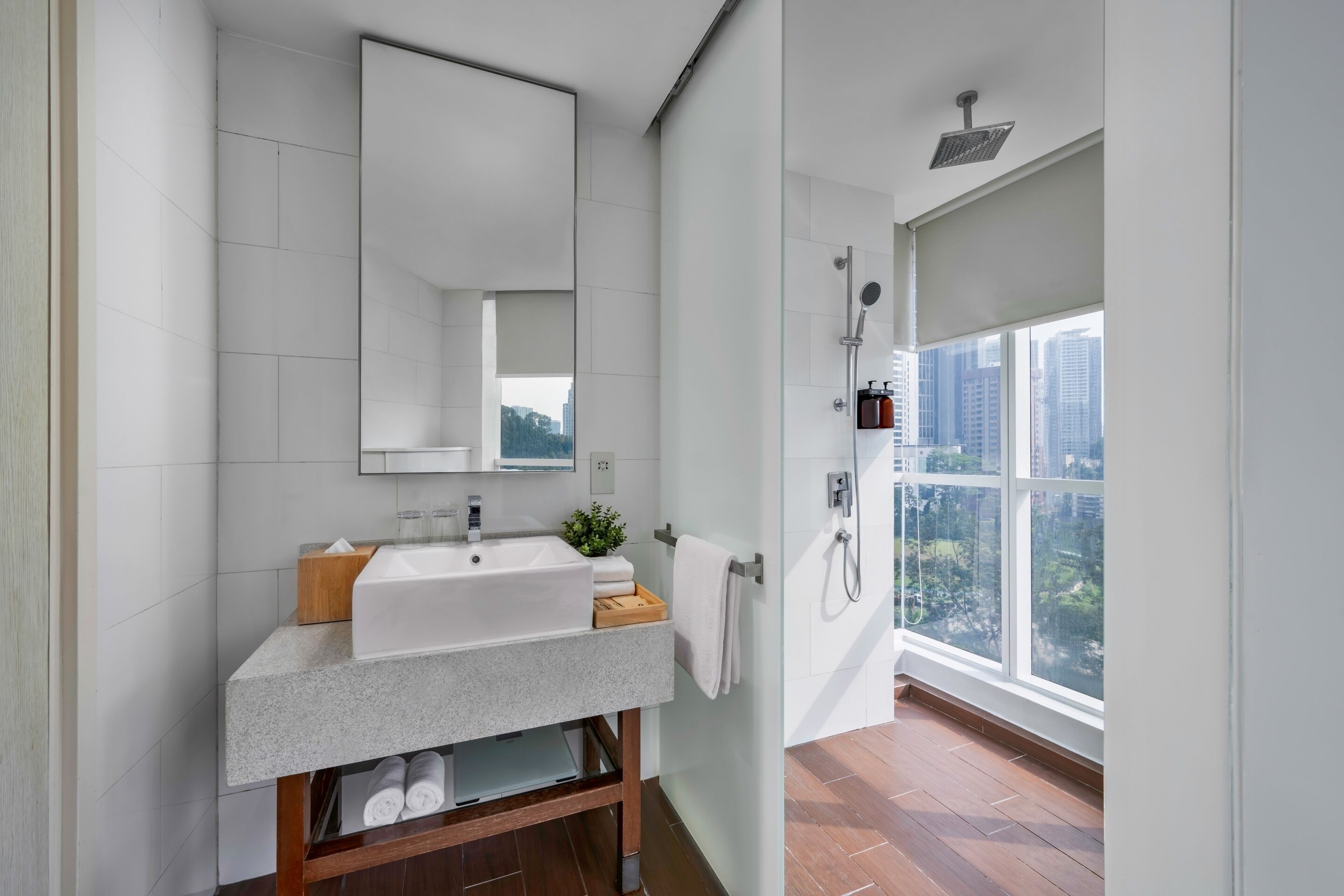 A modern bathroom with a large mirror, white sink, and a shower with a view of the city.