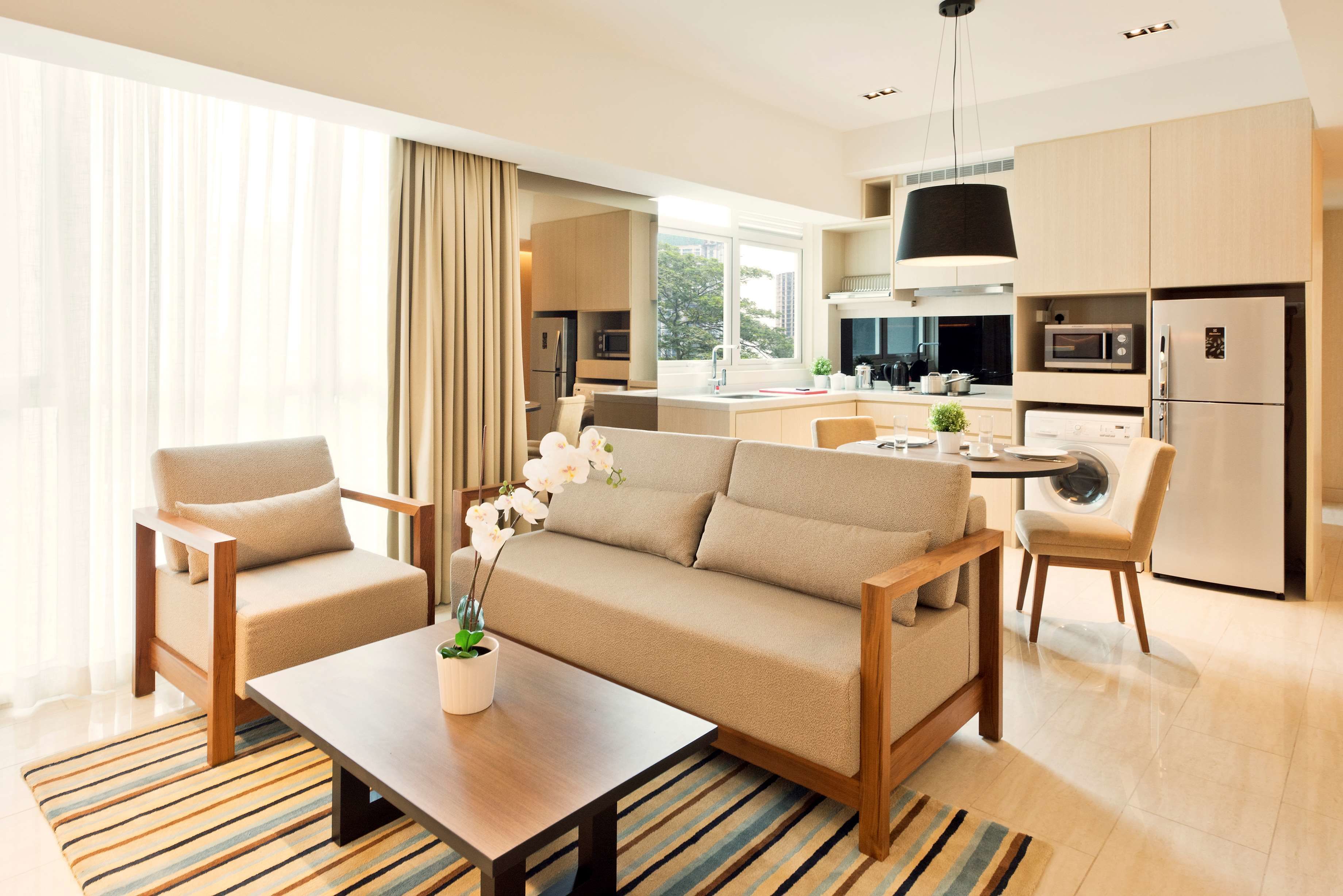 A modern hotel room with a sofa, two armchairs, a coffee table, a kitchen area with appliances, and a large window with curtains.