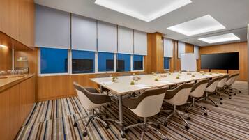 Un salón de conferencias con una larga mesa, sillas, una pizarra y un amplio ventanal con cortinas azules.