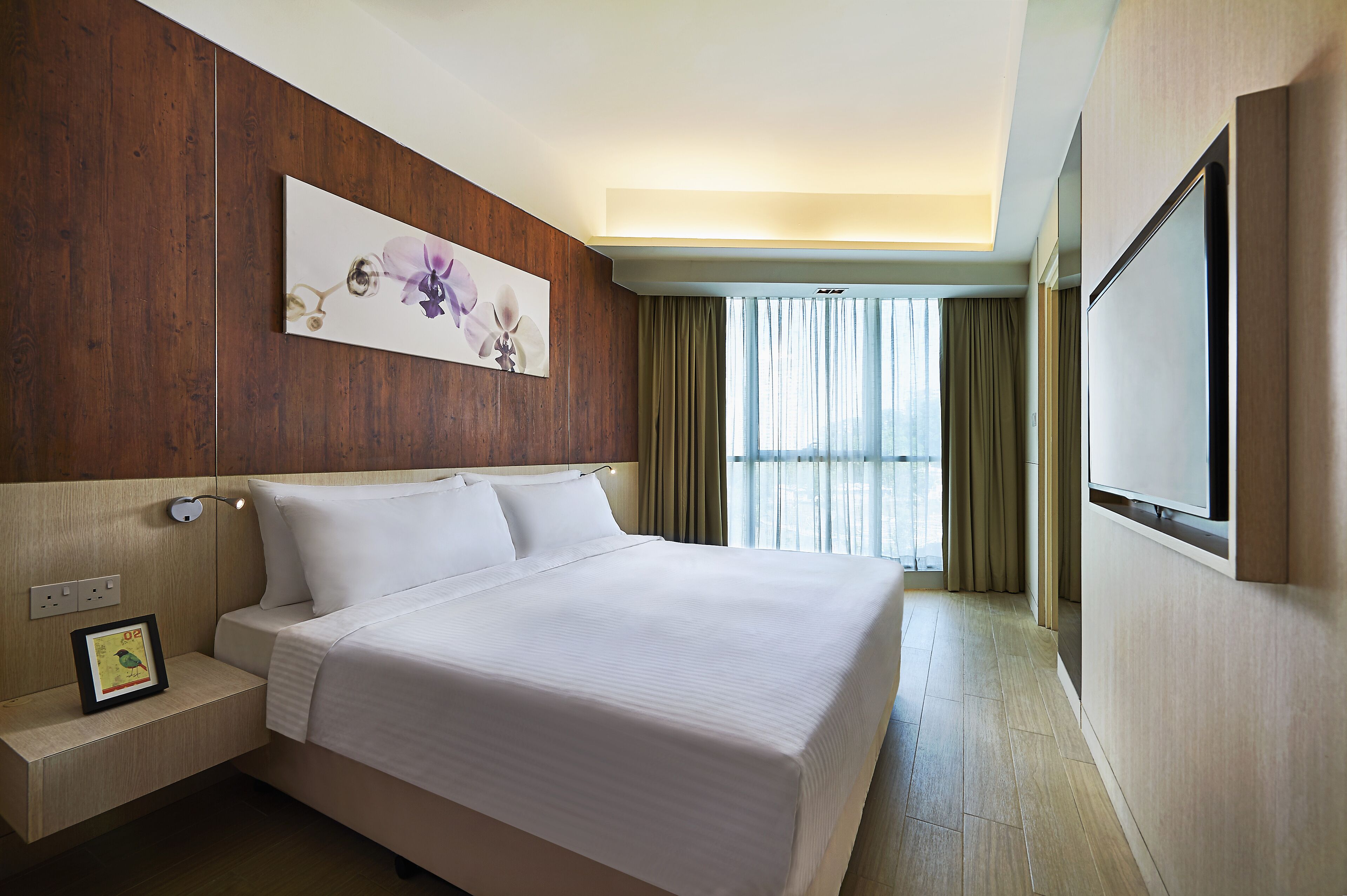 A hotel room with a large bed, a flat-screen TV mounted on the wall, a nightstand with a lamp, and a window with curtains.