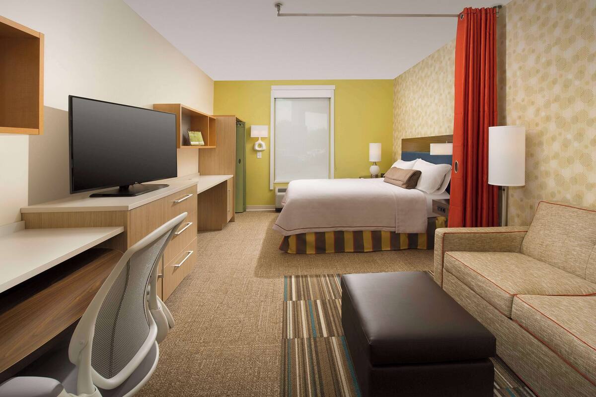 A hotel room with a bed, a desk with a computer, a TV, a sofa, and a chair.