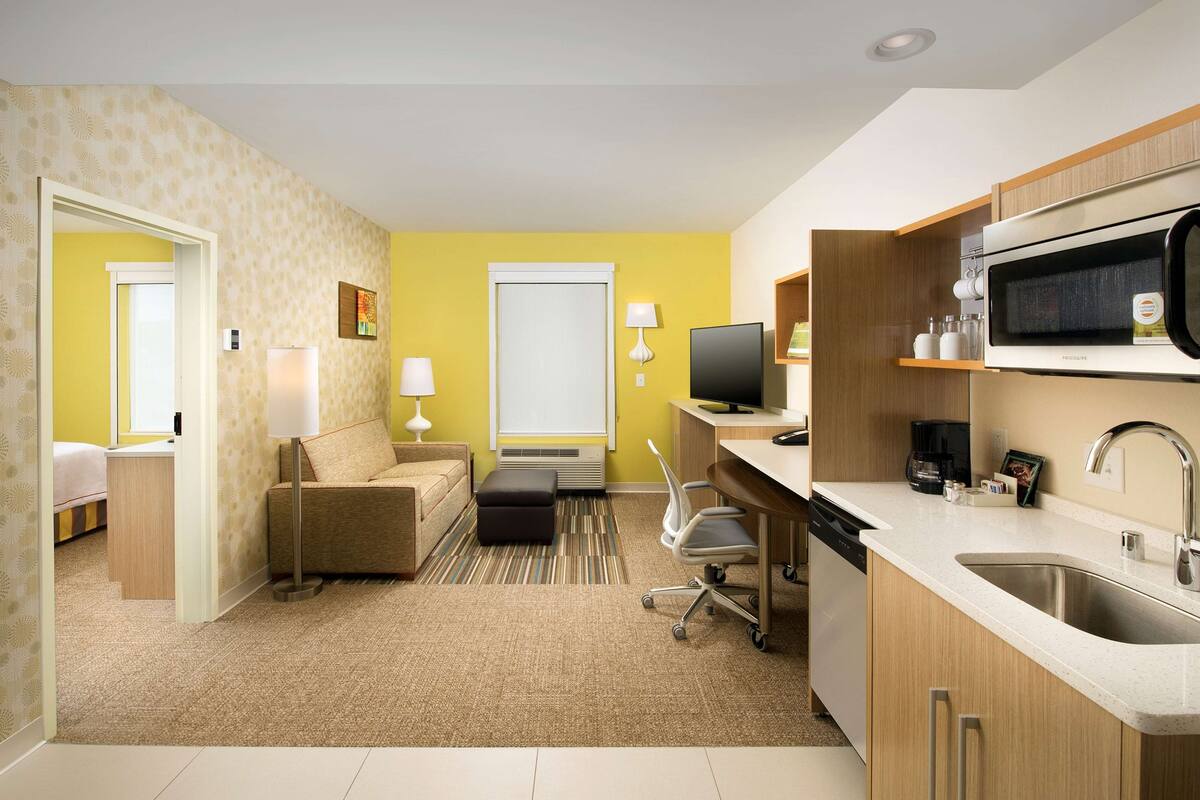 A compact hotel room with a kitchenette, a sofa, a dining area, and a separate bedroom.