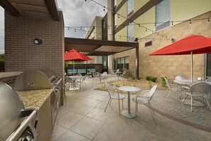 An outdoor patio with a barbecue grill, tables, and chairs, under a covered area with string lights. - Home2 Suites by Hilton Hattiesburg (Hattiesburg)