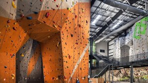 An indoor climbing facility with multiple climbing walls, a central wooden structure, and a mezzanine level with additional climbing routes.