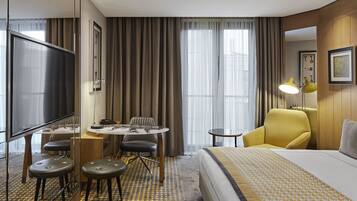 Habitación de hotel con cama, escritorio con silla, sillón amarillo, televisor y ventana con cortinas.