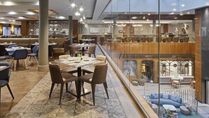 Un moderno vestíbulo de hotel con techo de cristal, zona de bar y área de descanso con mesas y sillas.