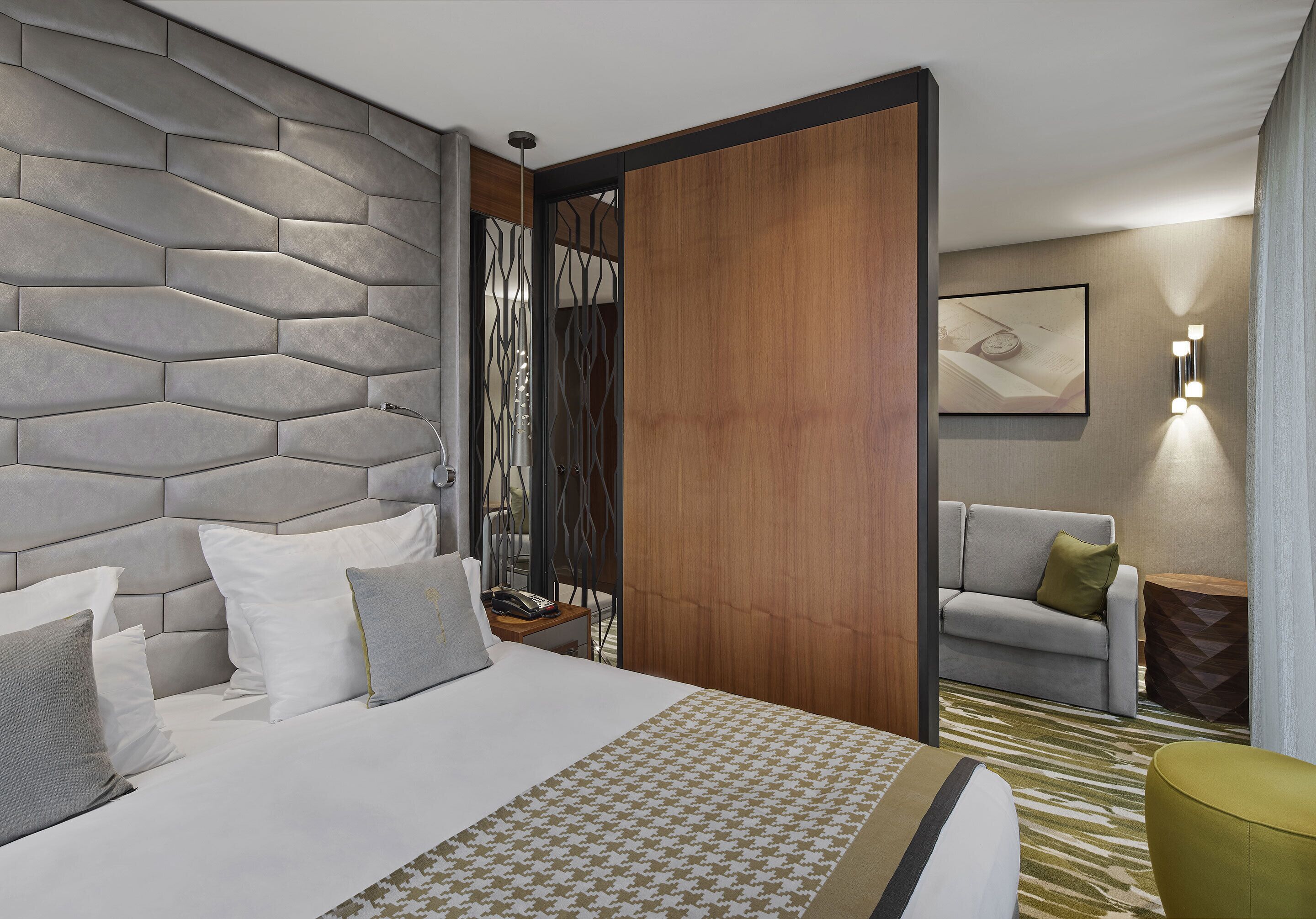 A modern hotel room with a large bed, a wooden partition, a grey sofa, and a patterned rug.