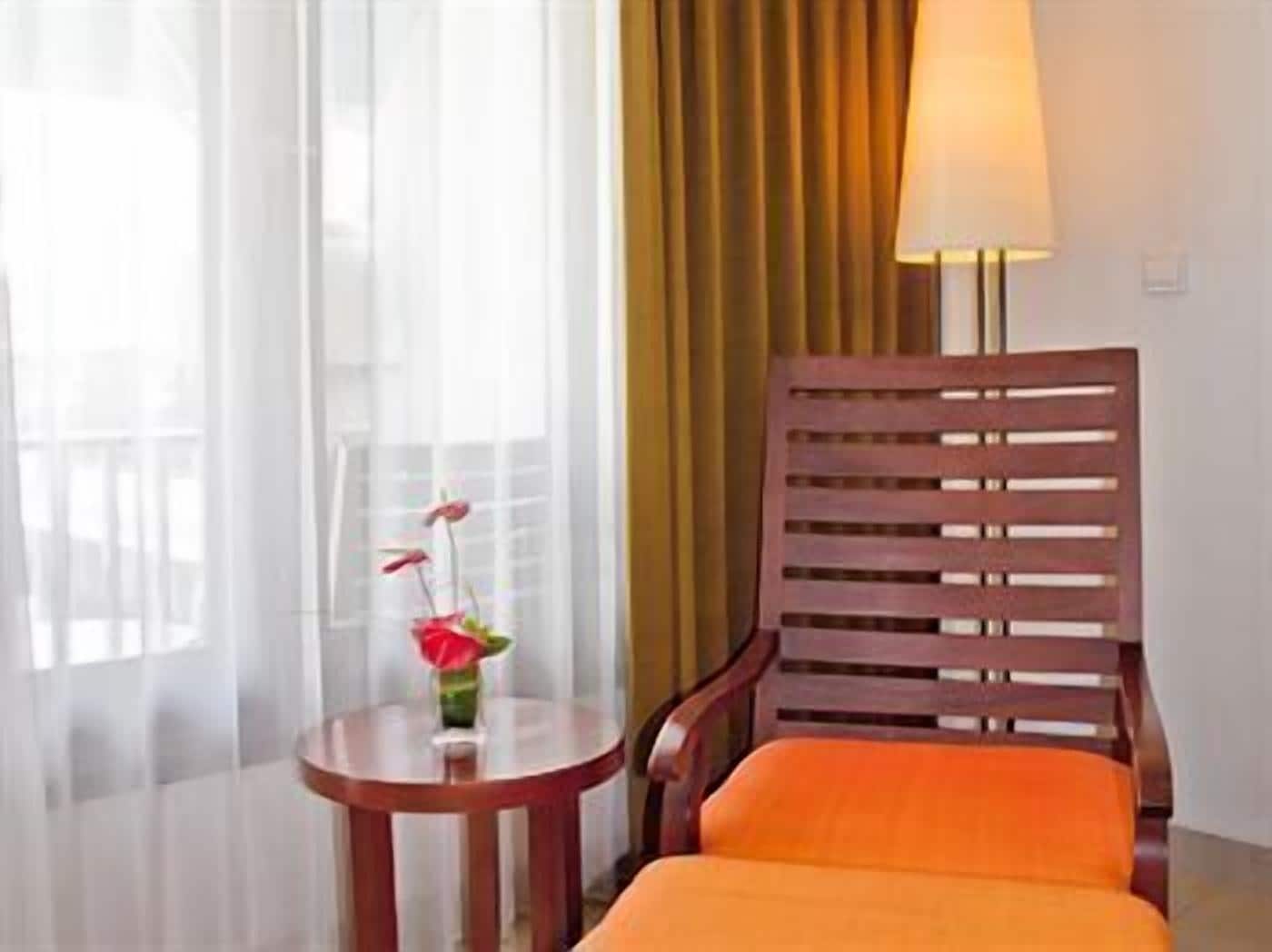 A hotel room with a wooden chair, a small round table with a vase of flowers, a lamp, and sheer curtains.