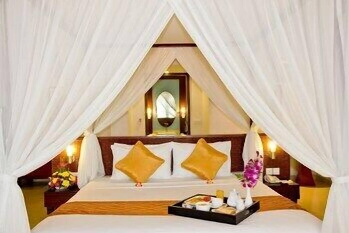 A bed with white linens, two gold pillows, a bedside table with a vase of flowers, and a tray with food and drinks.