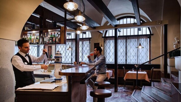 A bar area with a bartender preparing drinks, a customer seated at a table, and a staircase leading to an upper level. - Dell Angelo (Locarno)