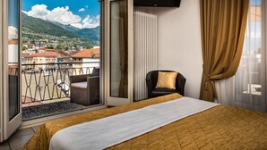 A hotel room with a bed, a chair, a TV, and a balcony with a view of mountains and buildings. - Dell Angelo (Locarno)