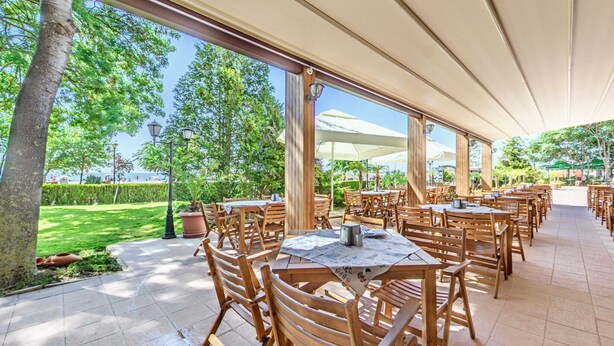 An outdoor dining area with wooden tables and chairs, a covered walkway, and a view of the sea. - Hotel Sentido Bellevue Beach Sunny Beach (Nessebar)